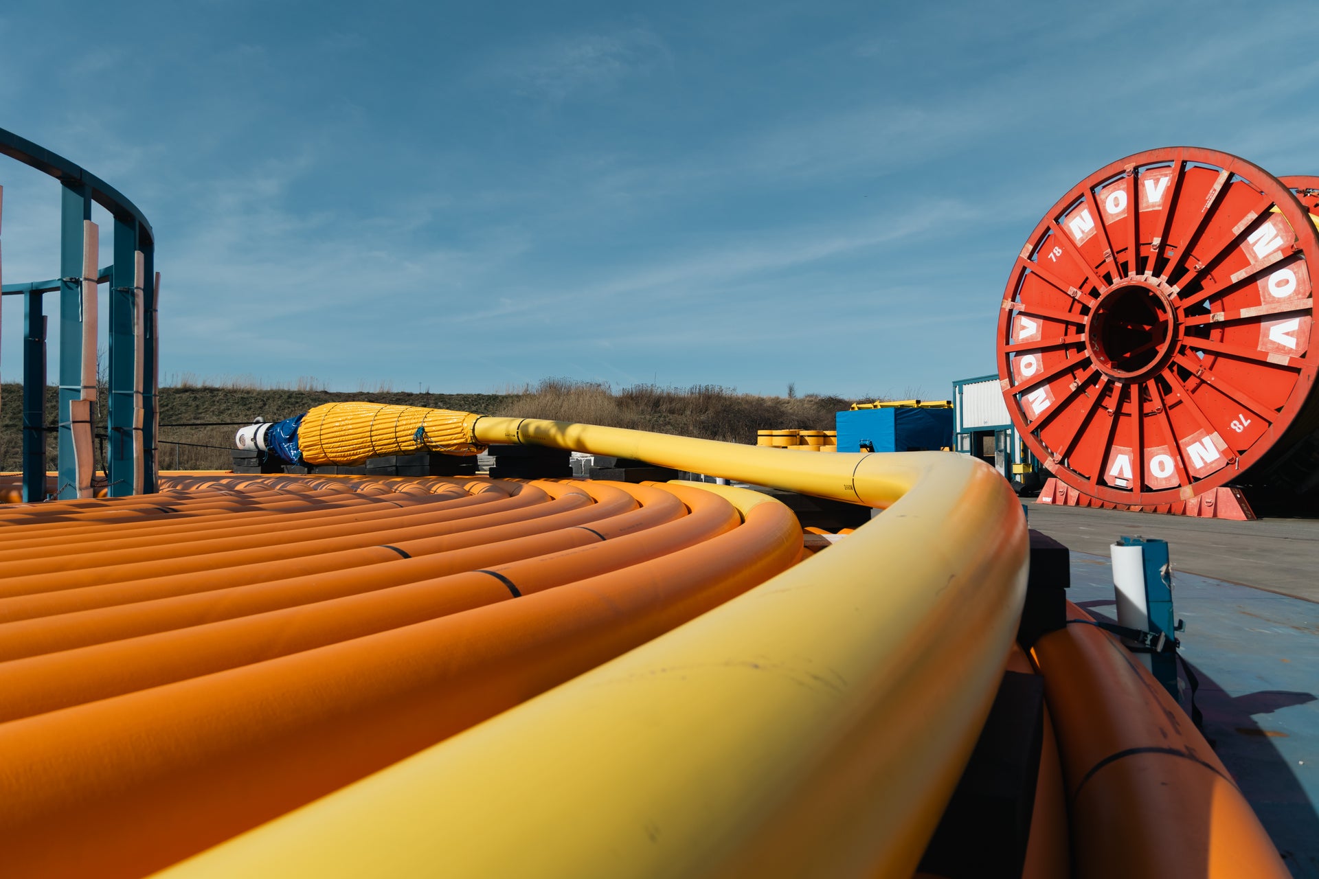 Large orange and yellow flexible offshore pipes and umbilicals are coiled in the foreground, leading toward a red industrial reel in an outdoor energy equipment storage yard under a clear blue sky.