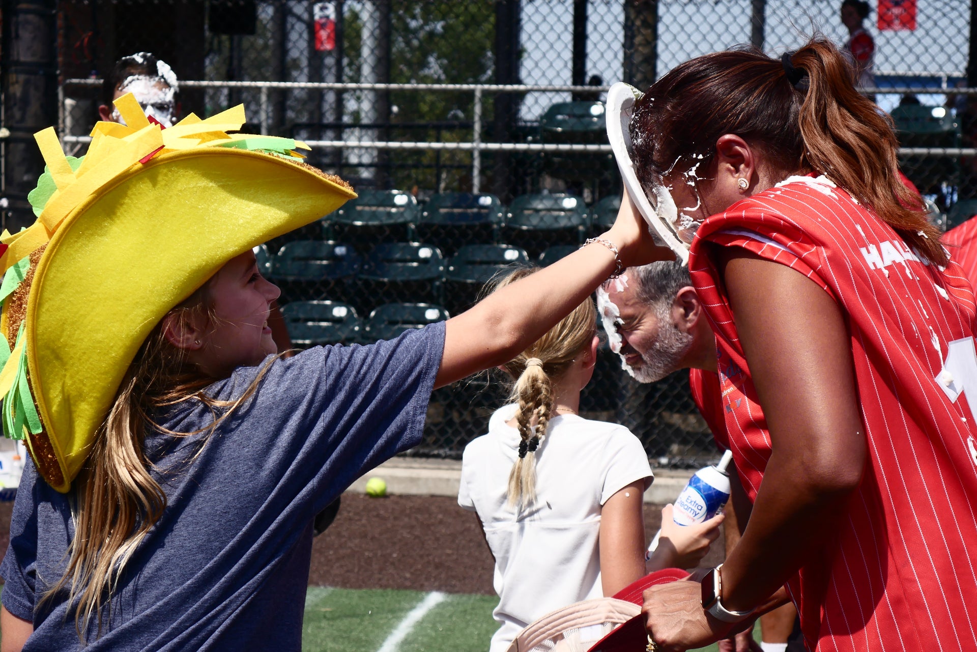 Woman getting pie thrown on her face by a kid wearing a taco hat during the Derricks and Diamonds softball charity tournament